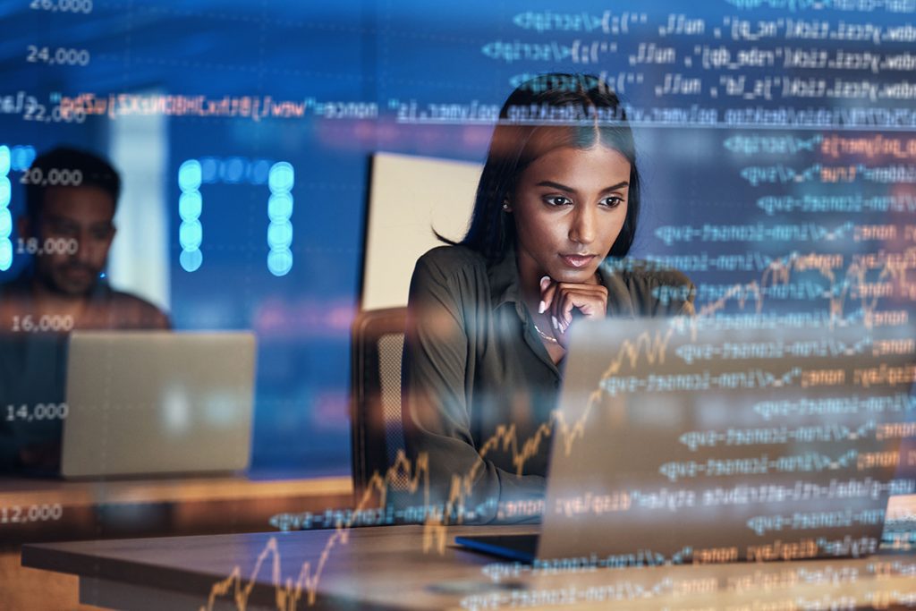 A woman looks intently at a laptop screen that is facing away from the camera. Behind the laptop, lines of code are displayed with a graph, showing what she is viewing.