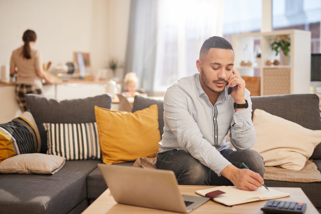 A man works at his living room coffee table with his wife and daughter in the background.
