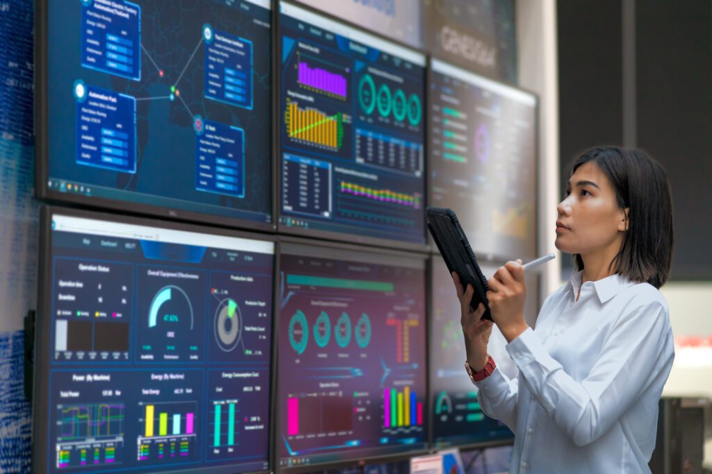A network admin holding a tablet looks at a large series of monitors that display data