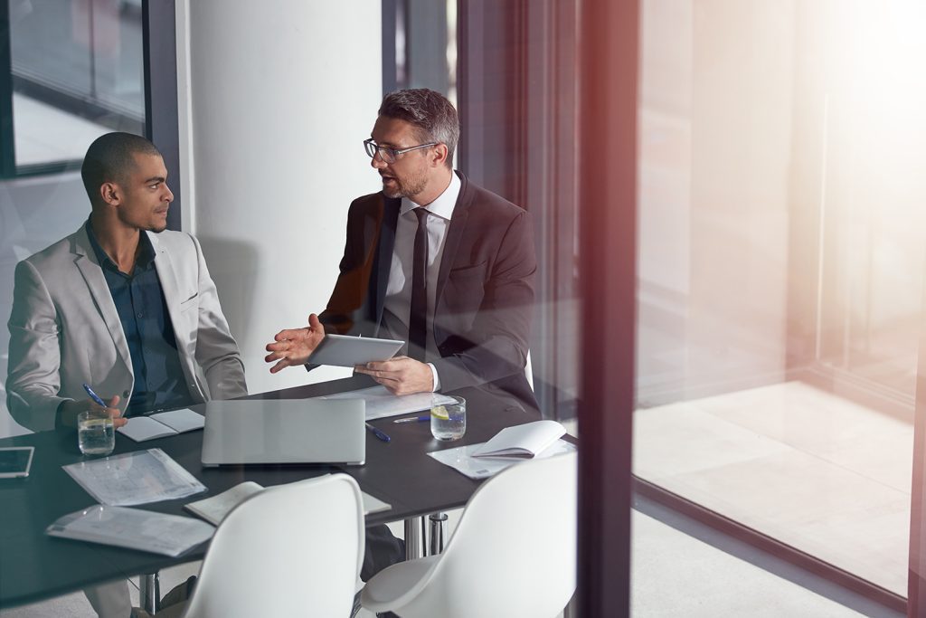 two men in suits sit in a conference room and discuss matters over a tablet, laptop, and papers.